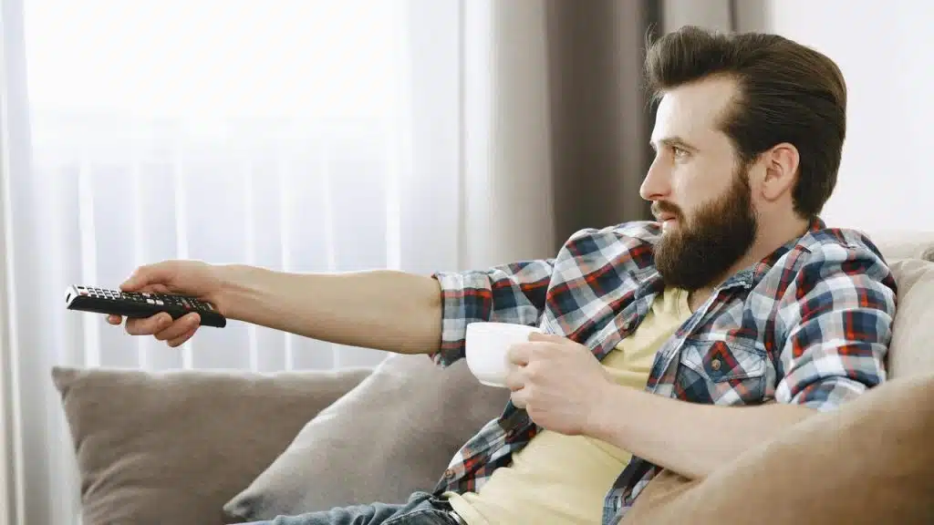 A man sitting on a couch holding a cup and using a remote control.