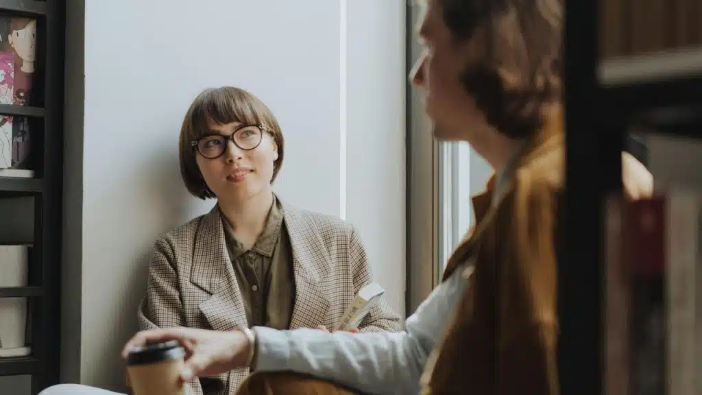 A woman wearing glasses holding a book while talking with another person indoors.