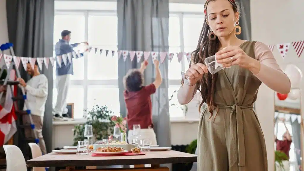 A woman arranging items on a table while others decorate a room in the background.