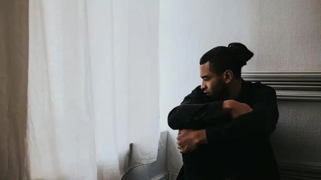A man sitting by a window, looking out with a pensive expression.
