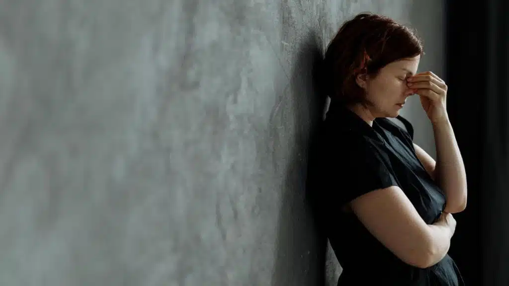 A stressed woman leaning against a wall with her hand on her face.