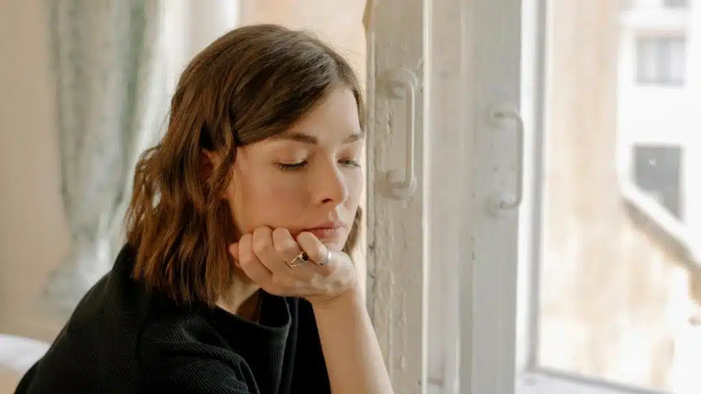 A pensive woman resting her chin on her hand by a window.