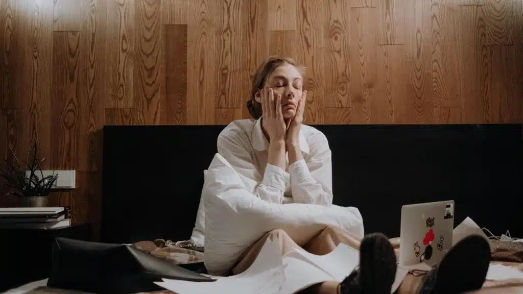 A tired person sitting on a bed with hands on their face.