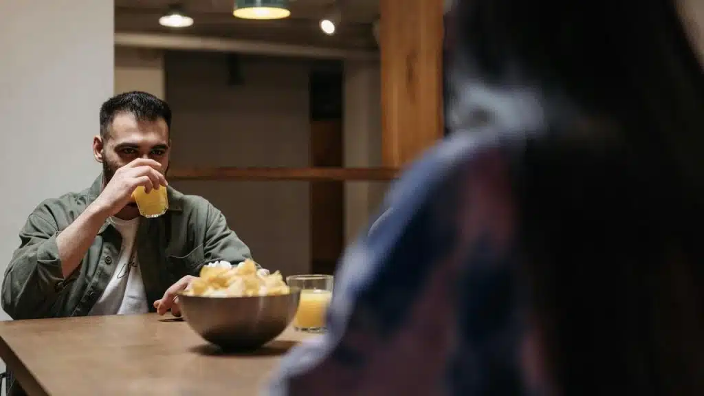 A man drinking juice while sitting at a table across from another person.