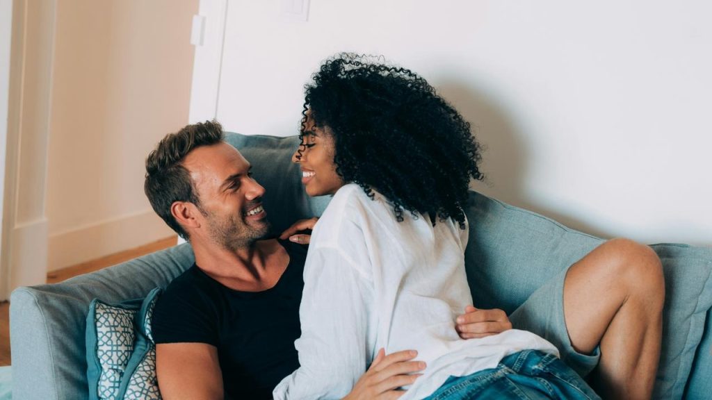 A couple relaxing on a couch, smiling closely at each other indoors.