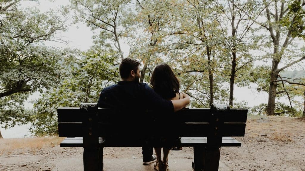 A couple sitting close together on a park bench, looking out at trees and water.