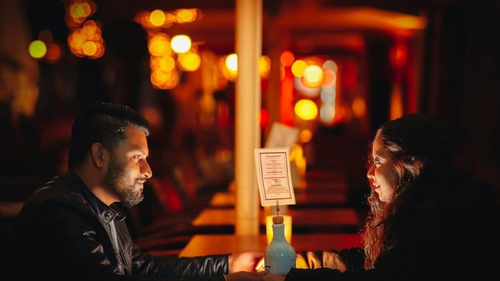 A couple holding hands across a table at a warmly lit restaurant at night.