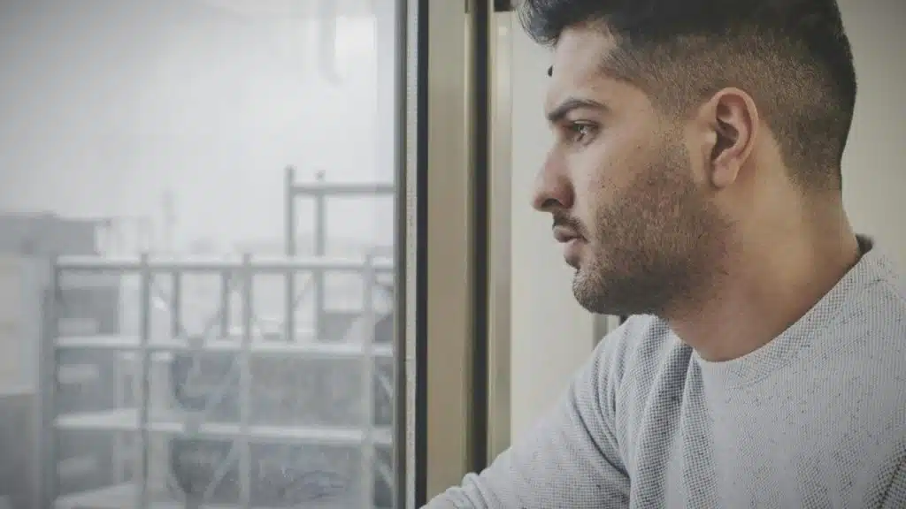 A man gazes out a window with an industrial scene visible outside.