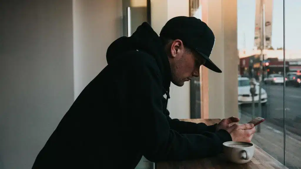 A man in a hoodie sitting by a window, checking his phone with a cup of coffee nearby.
