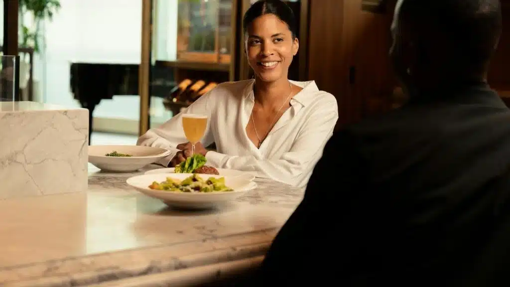 A woman smiling at a dining companion across a marble table with plated food and a drink.