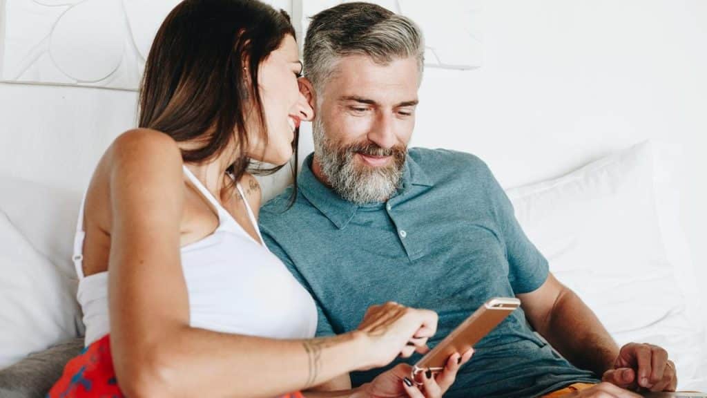 A couple relaxing on a bed while smiling and looking at a smartphone together.