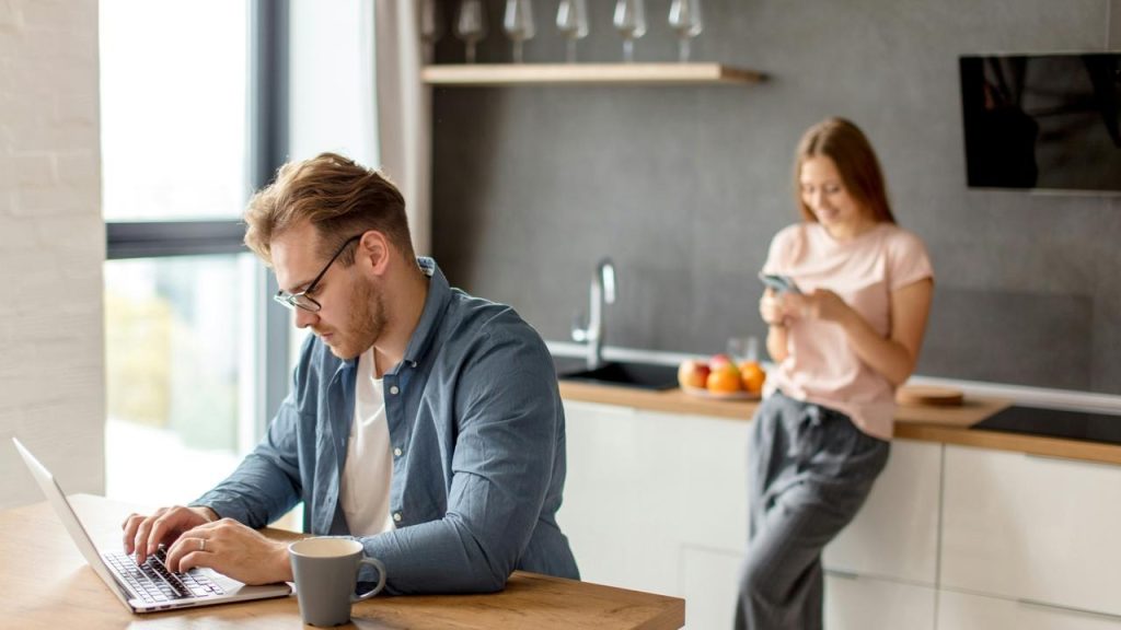 A man working on a laptop at a table while a woman looks at her phone in the background.