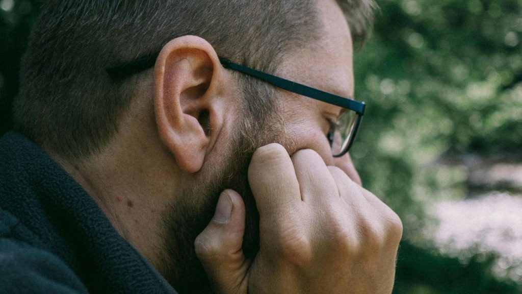 A man wearing glasses touching his beard outdoors.