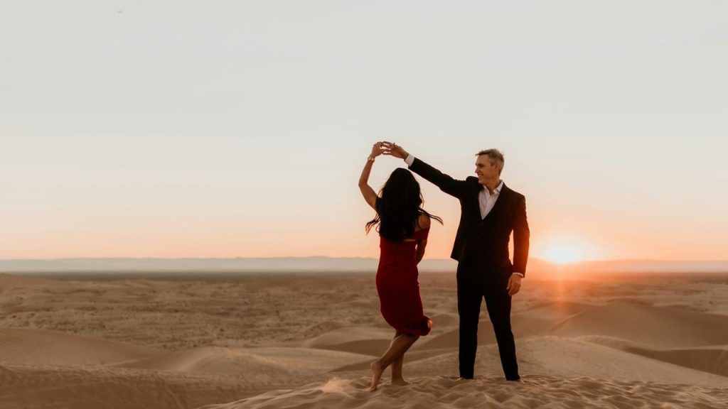 A couple dancing together on sand dunes at sunset, silhouetted against the horizon.