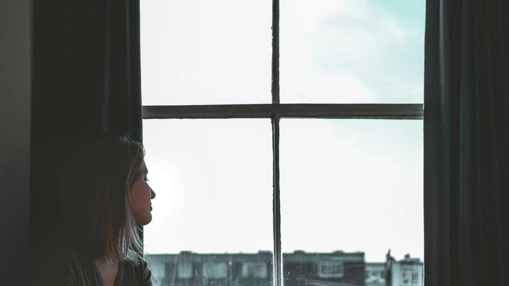 A woman in profile gazing out a large window with curtains and city buildings beyond.
