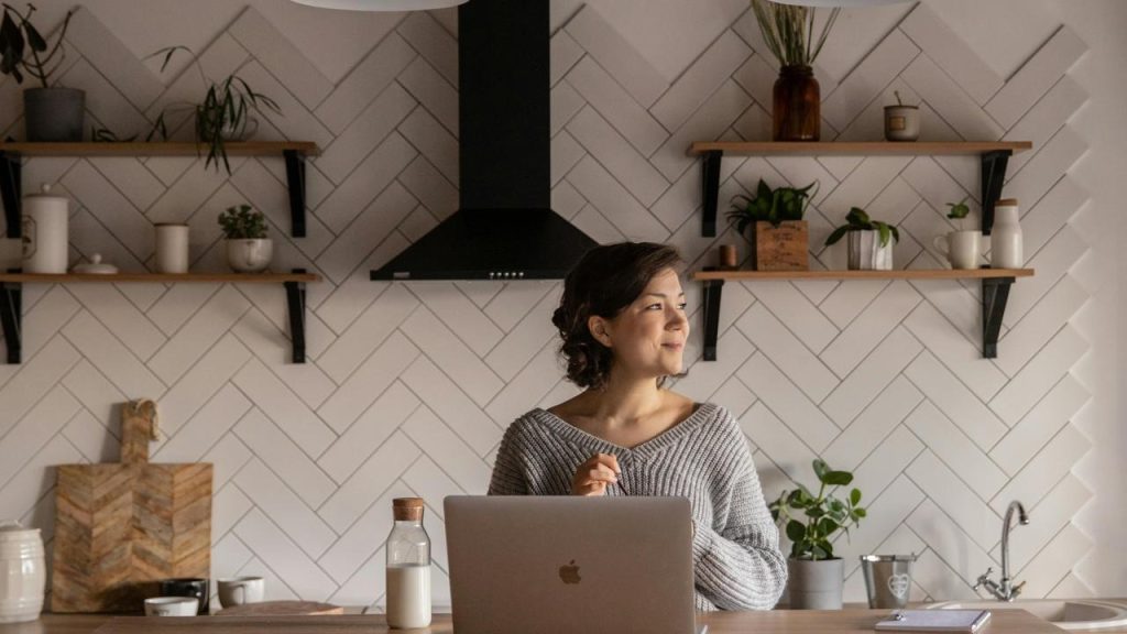 A woman standing in a modern kitchen with a laptop, looking thoughtfully to the side.