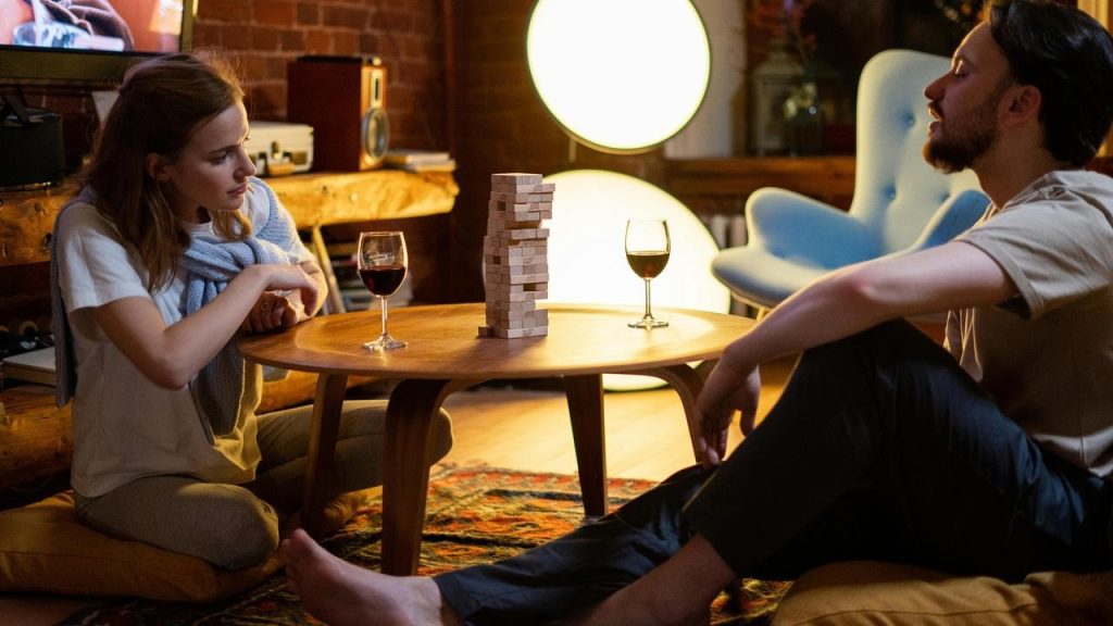 A couple sitting on the floor playing a block-stacking game with wine glasses on a small table.