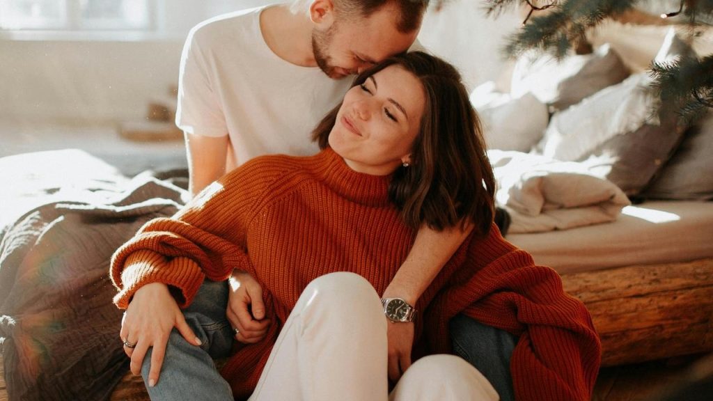 A couple cuddling indoors, with one person embracing the other from behind.