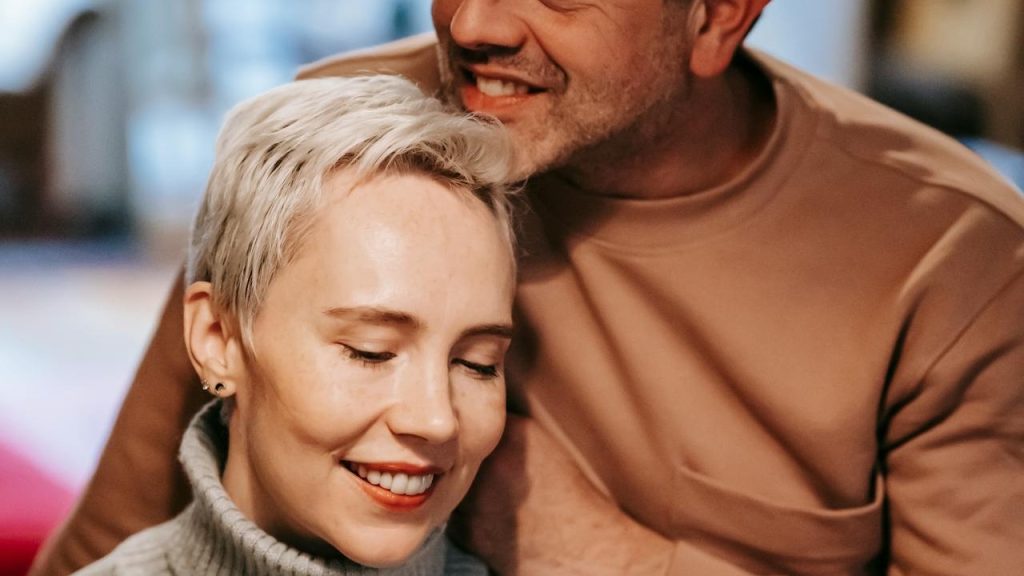 A close-up of a woman with short blonde hair smiling while a man leans in close to her, his chin resting near the top of her head.