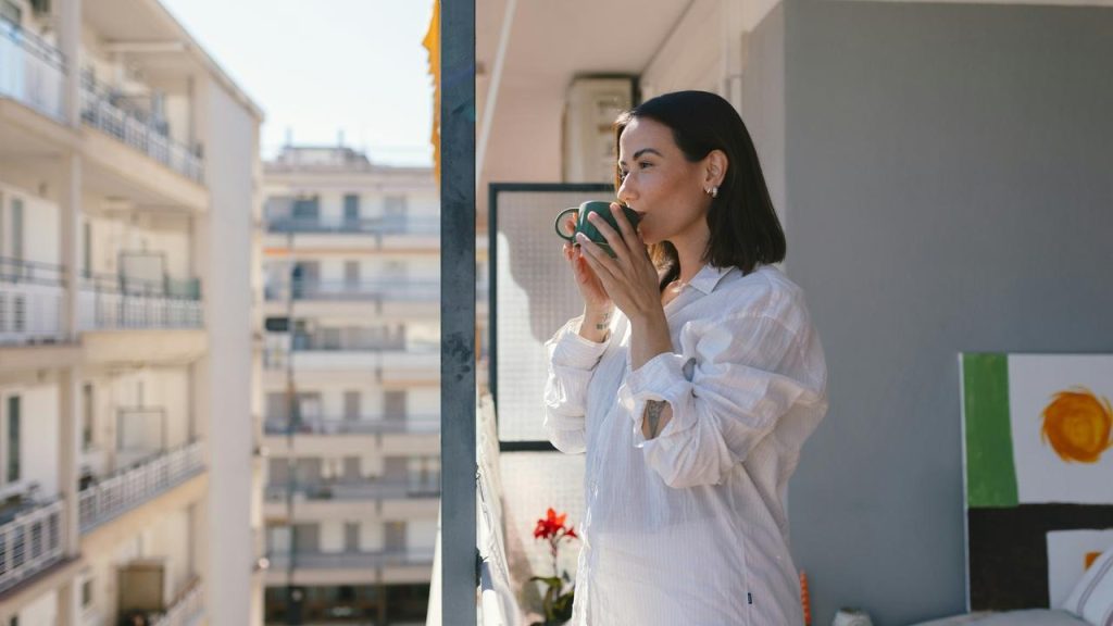 A woman standing on a balcony, sipping from a cup while looking at the city outside.