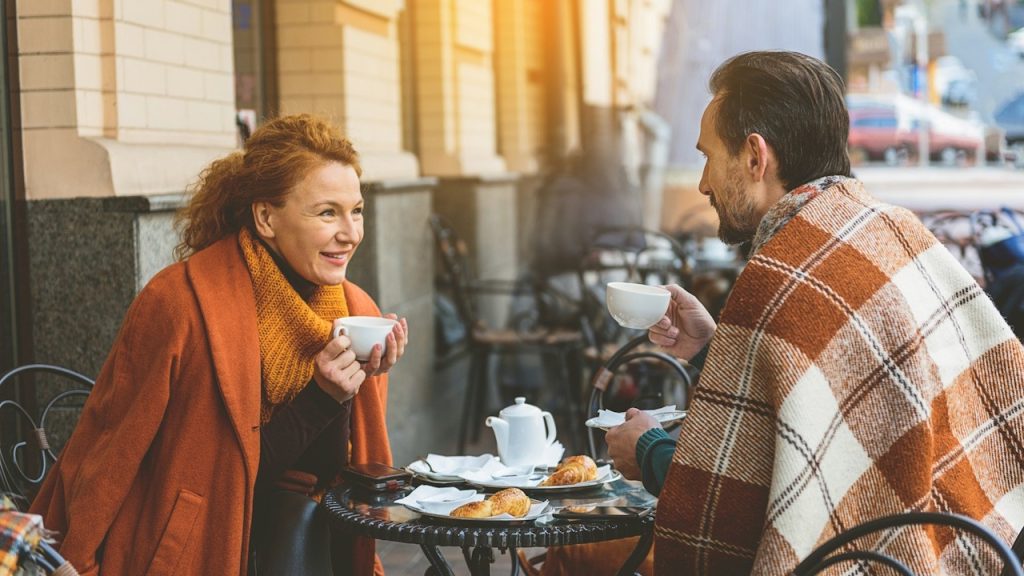 A man and woman enjoy coffee and pastries at an outdoor café table, with the man wrapped in a plaid blanket.