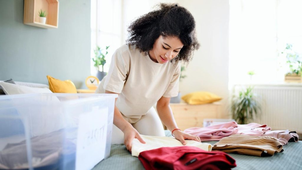 A woman folding clothes on a bed next to a plastic storage bin.