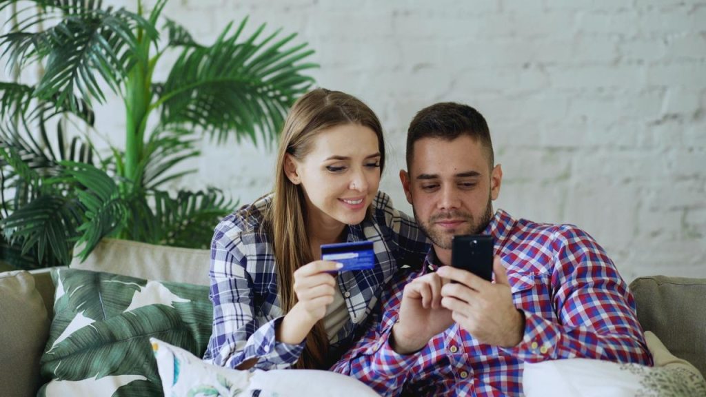 A smiling couple sitting on a couch, shopping online with a credit card and smartphone.