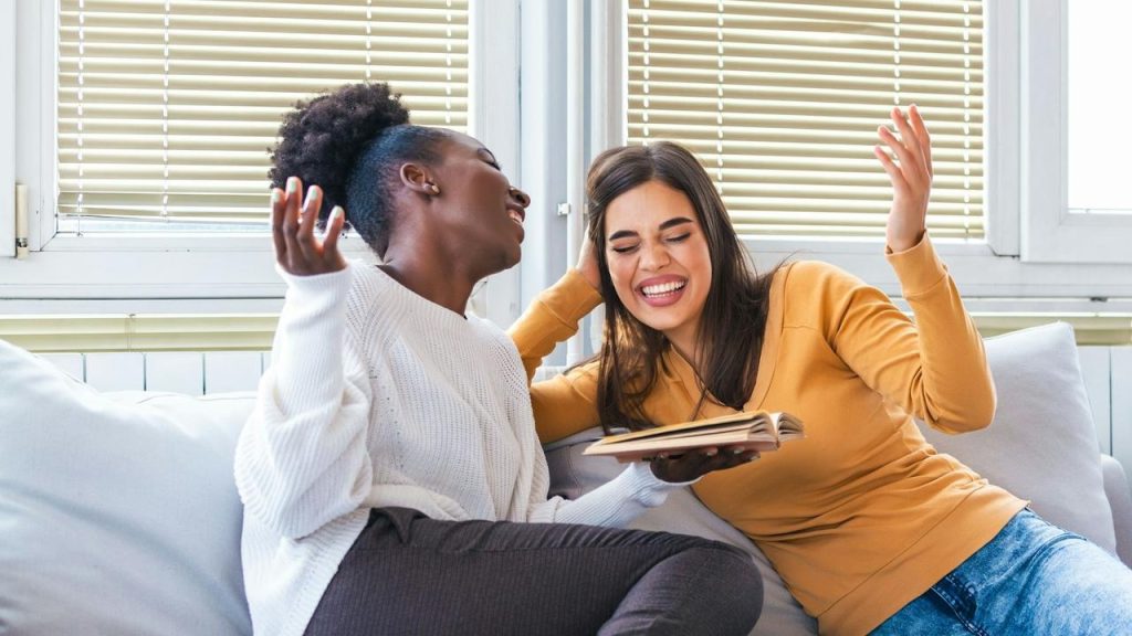 A pair of women laughing together on a couch with a book