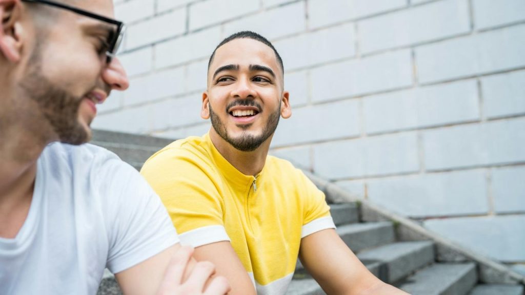 A man in a yellow shirt smiling while talking to a friend.