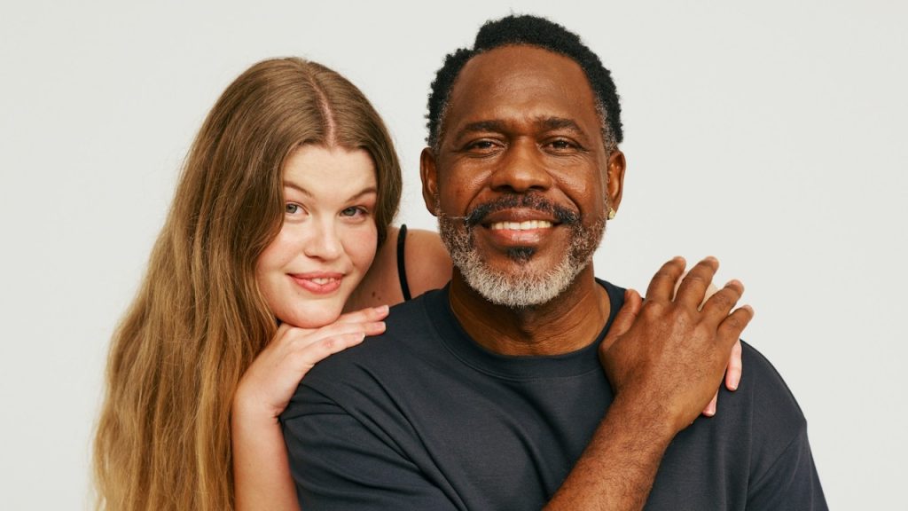 A smiling man and woman pose together against a plain white background, with the woman leaning her chin on her hands resting on the man's shoulders.