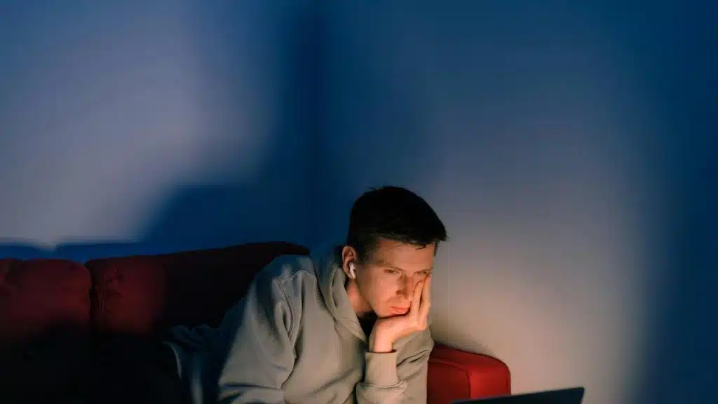 A tired person leaning on their hand while looking at a laptop on a couch in dim lighting.