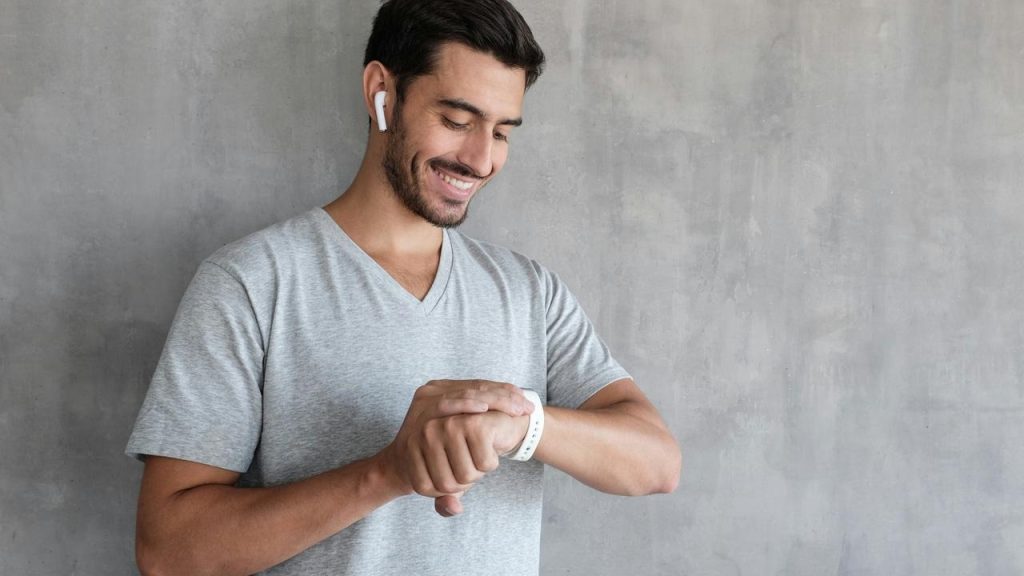 A smiling man wearing earbuds checking his smartwatch against a gray wall.