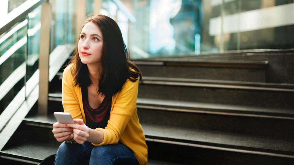 A woman sitting on stairs holding a phone and looking thoughtfully into the distance.