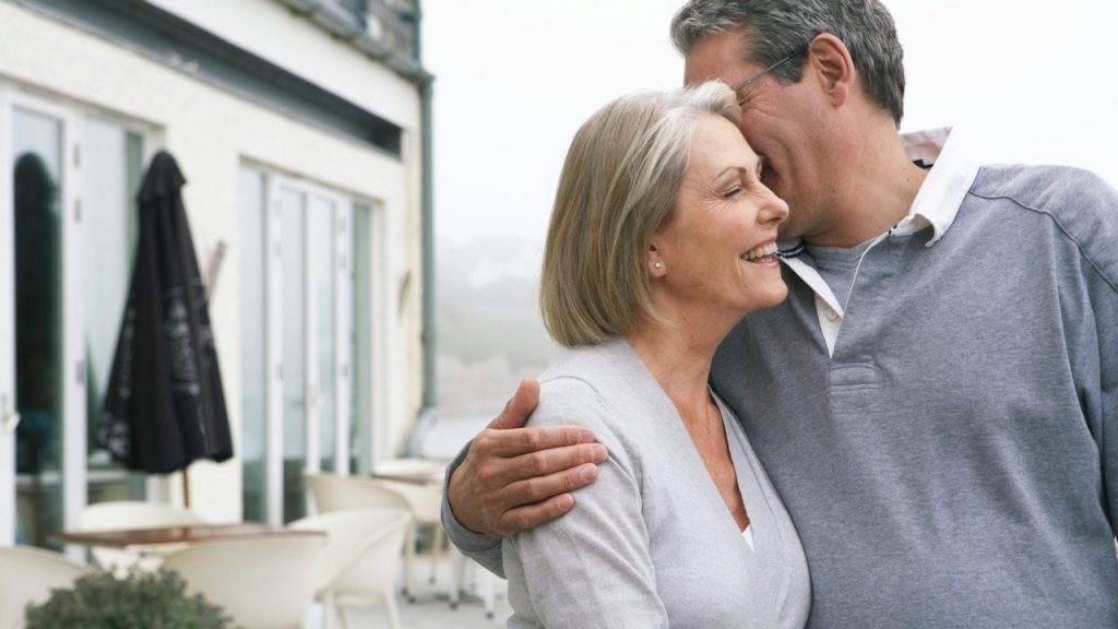 A smiling mature couple embracing warmly on an outdoor terrace, with the man leaning in toward the laughing woman.