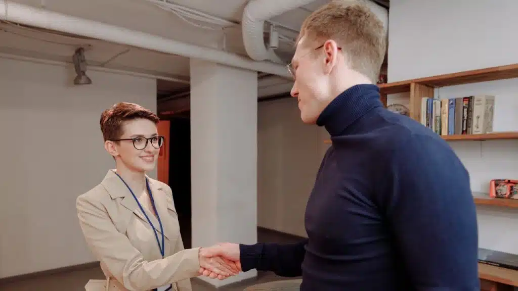 A woman in a beige coat and glasses smiling while shaking hands with a man in a navy blue turtleneck in an office or library setting.