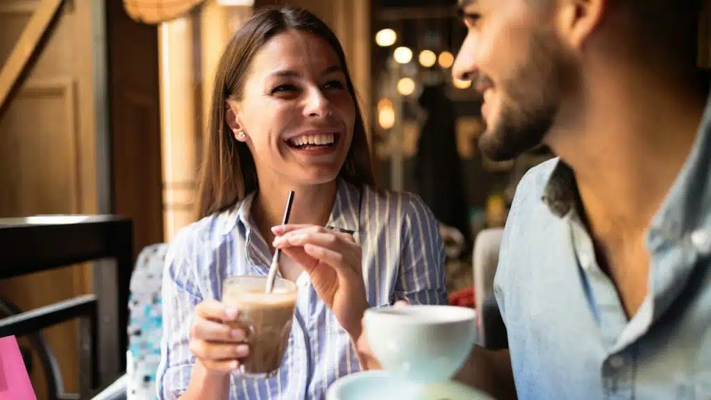 A woman laughing and holding a drink with a straw while sitting across from a man holding a coffee cup in a brightly lit cafe.