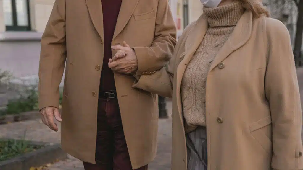 A man and a woman wearing matching tan overcoats hold hands while walking together on a brick-paved sidewalk.