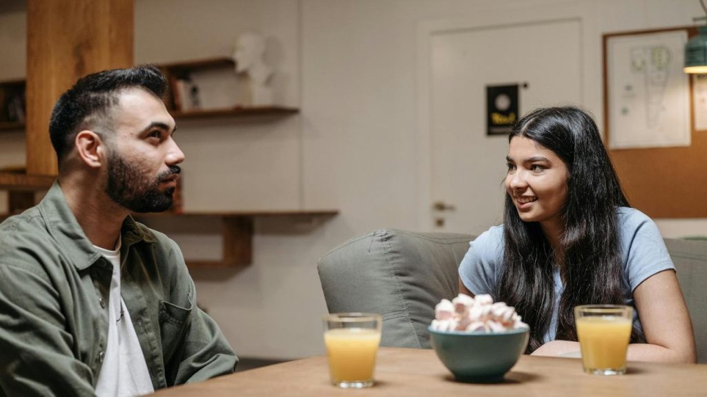 A man and a woman sitting at a table, talking with drinks and a bowl of snacks between them.