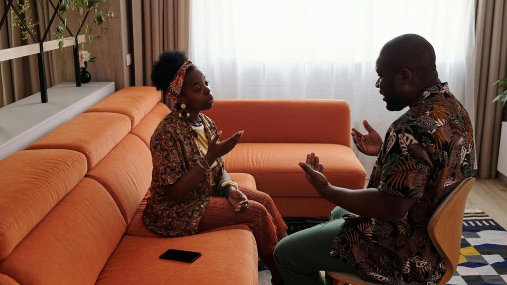 A man and woman talking and gesturing while sitting on an orange couch.