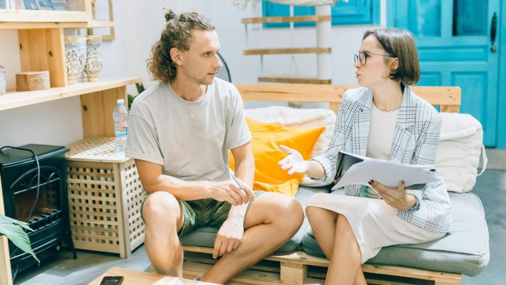 A woman holding papers talking to a man sitting on a couch.