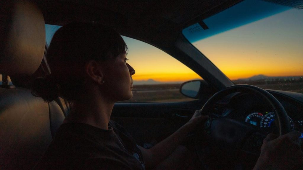 A woman driving a car at sunset, looking ahead through the windshield.