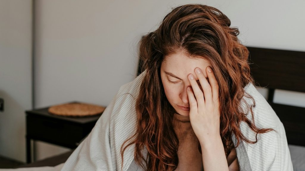 A woman looking distressed and holding her face with her hand as she sits on a bed.