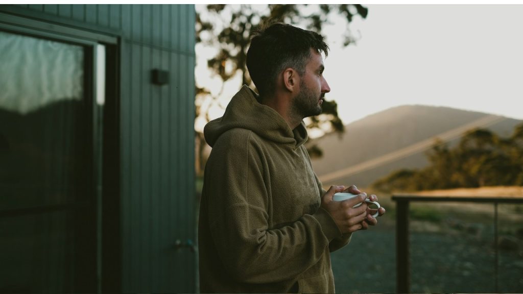 A man is holding a coffee cup, standing outside his home and looking at the scenery.
