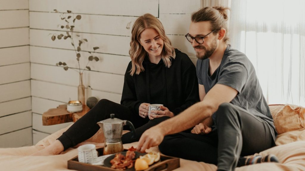A man serving breakfast to his wife while they are sitting in the bedroom.