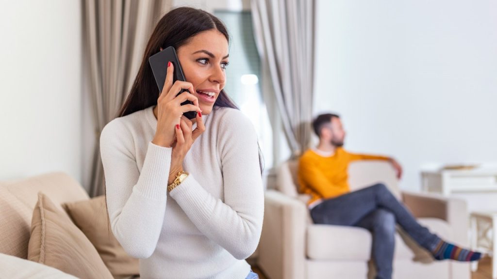 A woman is talking on the phone while her husband is sitting in the background.