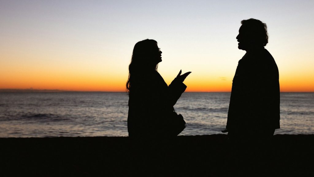 A silhouette of a couple arguing in front of the ocean on a beach at sunset.