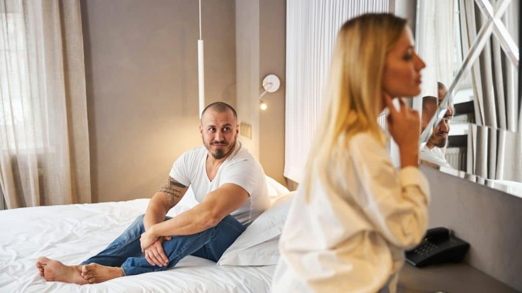 A handsome, serene Caucasian man is seated in bed, looking at a serious blonde woman.