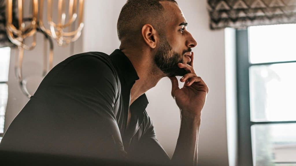 A thoughtful man in profile resting his chin on his hand by a window.
