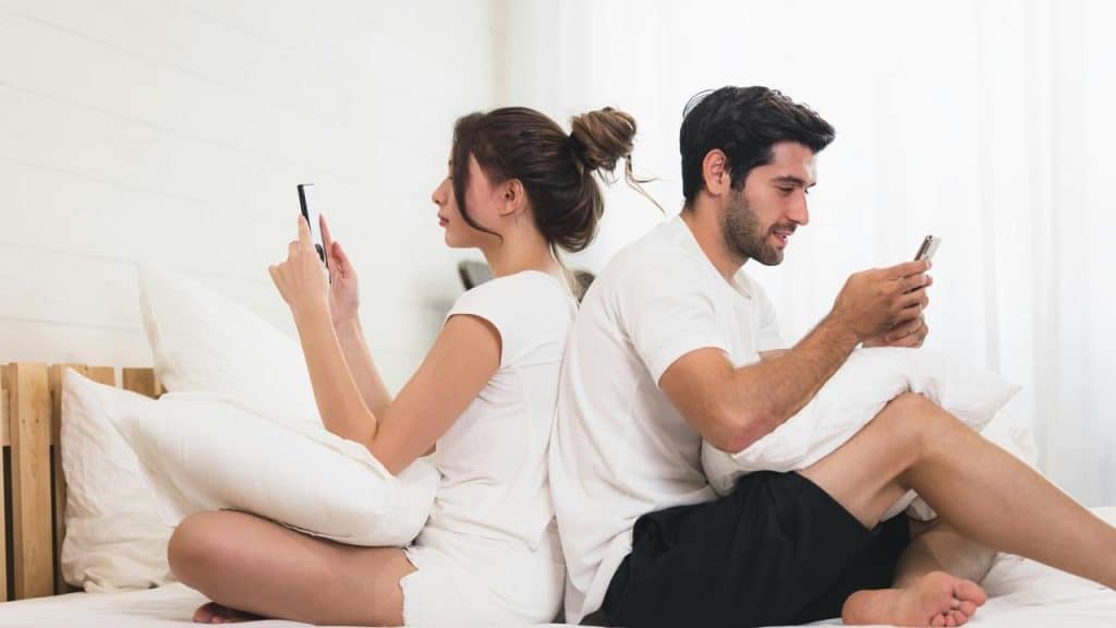 A couple sitting back to back on a bed, each focused on their phone.