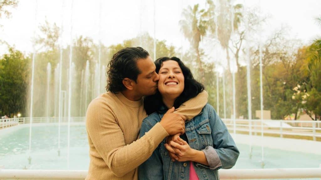A couple hugging by a park fountain, smiling.